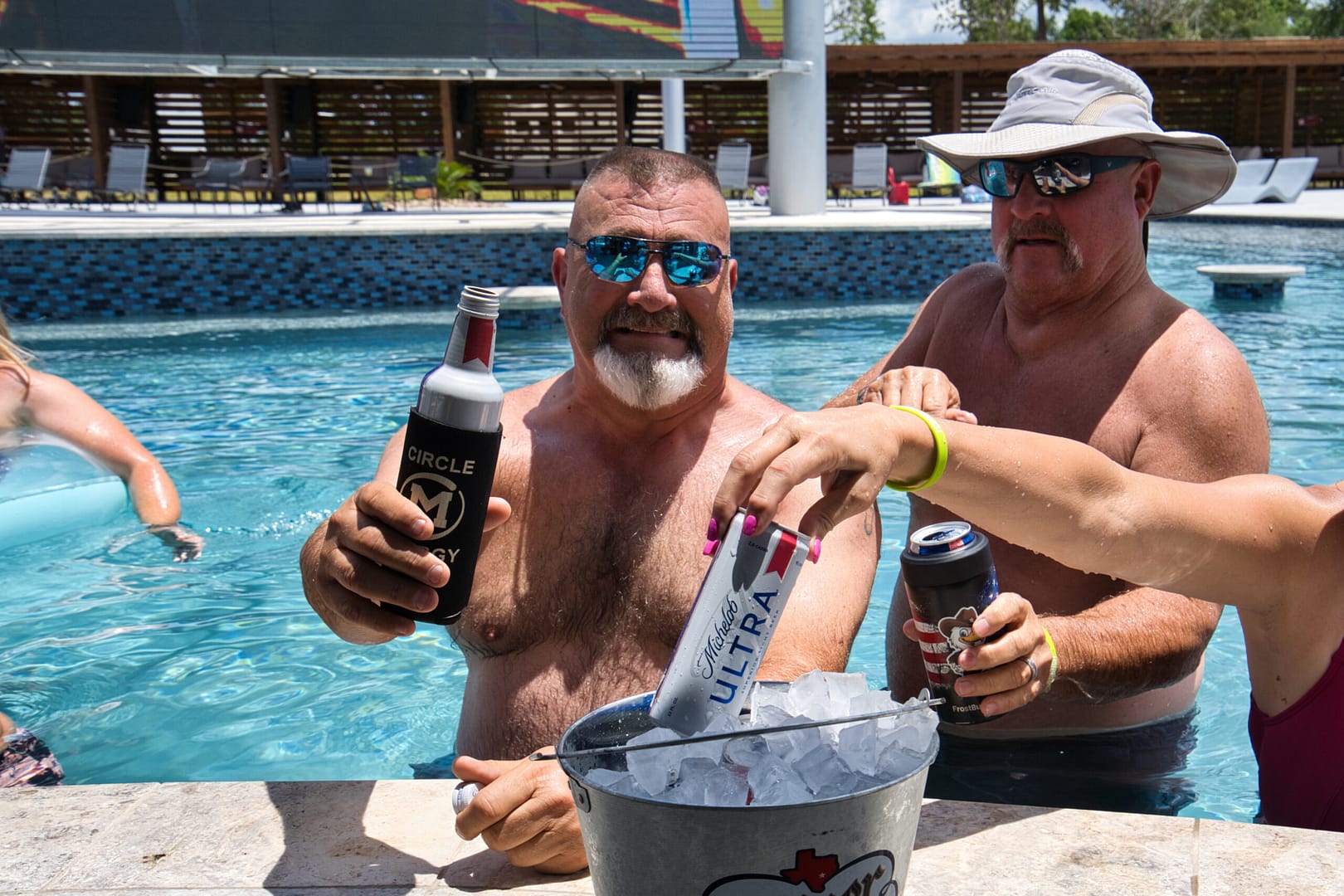 Two men in a pool hold drinks while a woman’s hand reaches for a can from an ice bucket. The sun is shining and they appear relaxed and smiling.