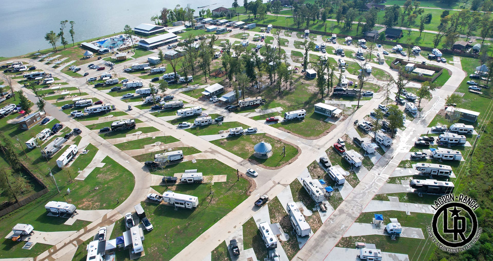 Aerial view of a large RV park with numerous RVs parked on paved lots, trees scattered throughout, a pool area, and a nearby lake.