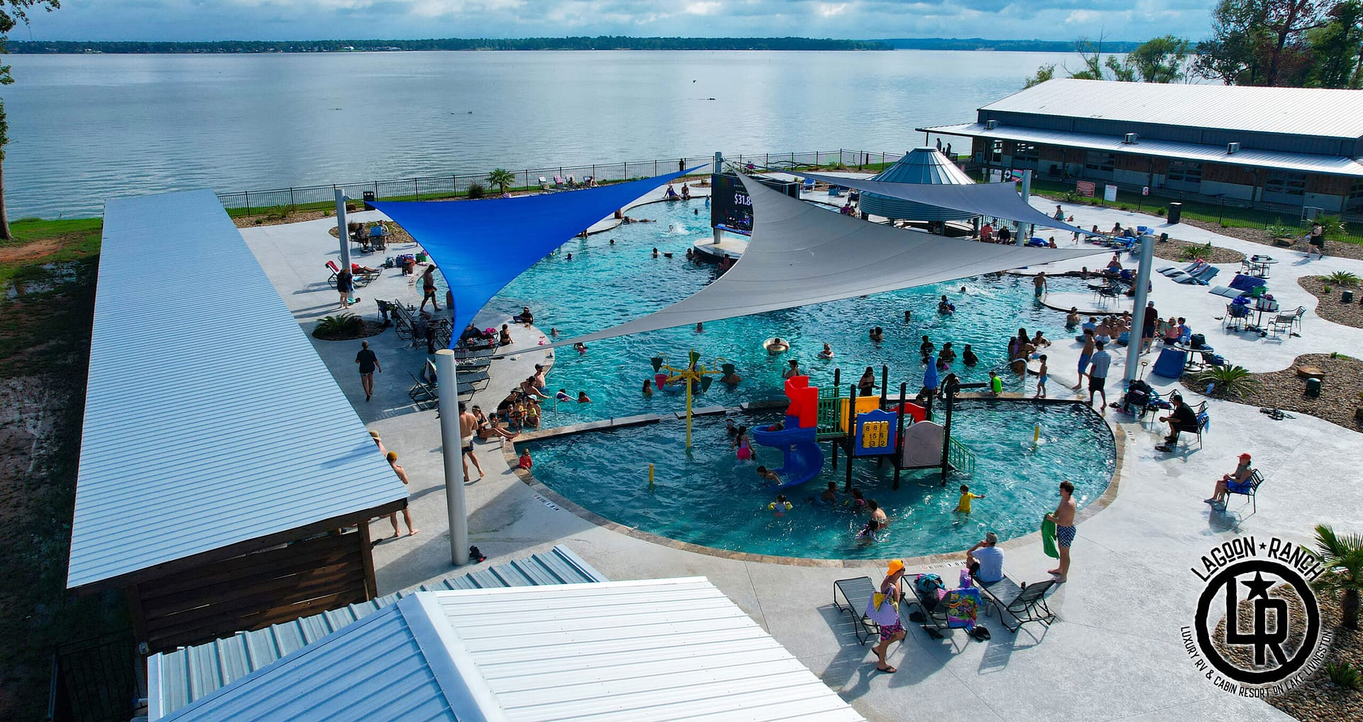 A public pool with shade sails, people swimming and relaxing, a children's play area, and seating around the pool, located next to a large lake under a partly cloudy sky.