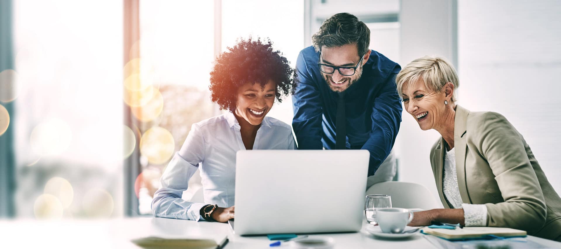 Three business professionals, two women and one man, sit together at a desk, smiling and looking at a laptop screen in a bright, modern office setting with papers and a glass of water nearby.