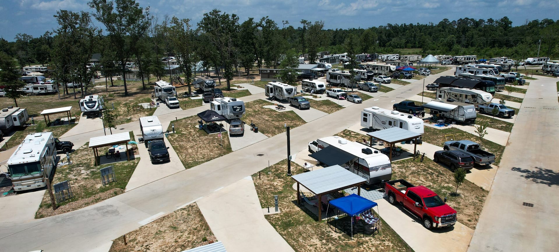 Aerial view of an RV park with multiple RVs and trucks parked in designated spots, surrounded by trees and paved roads under a partly cloudy sky.