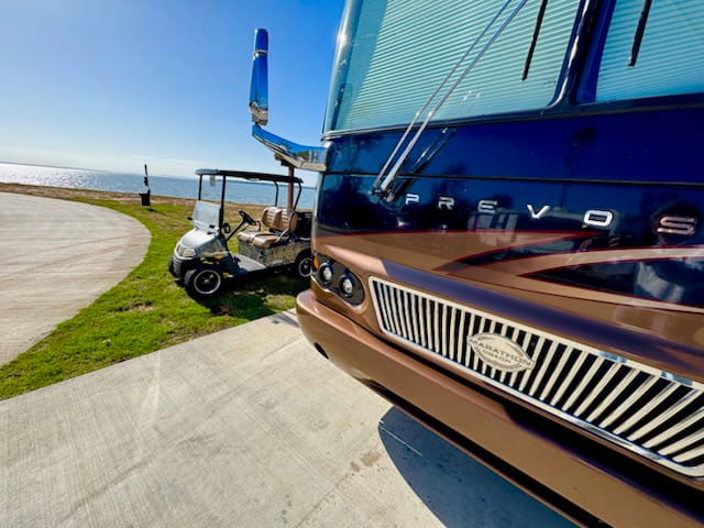 A large RV and a golf cart are parked on a concrete pad near a grassy area by the water under a clear blue sky.