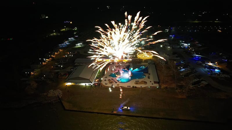 Aerial view of fireworks exploding over a campsite with a pool at night, surrounded by RVs and illuminated buildings.
