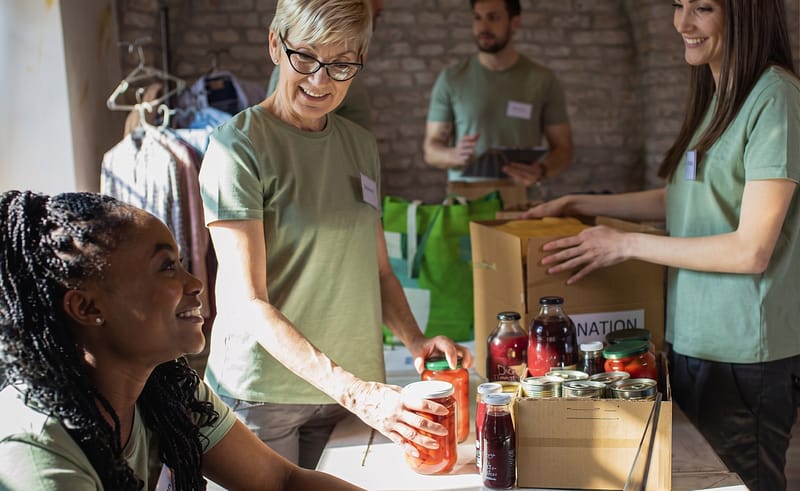 Four people in matching shirts sort food and canned goods at a donation table while one woman works on a laptop.