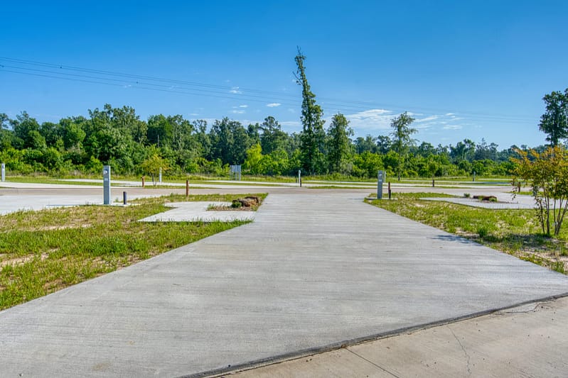 A wide, empty concrete lot with several utility hookups, surrounded by grass and trees under a clear blue sky. The area appears to be part of an RV park or campground.