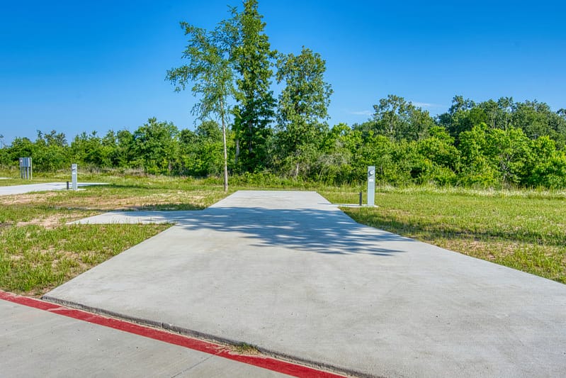 Paved RV parking pad with utility hookups, surrounded by grass and trees under a clear blue sky.