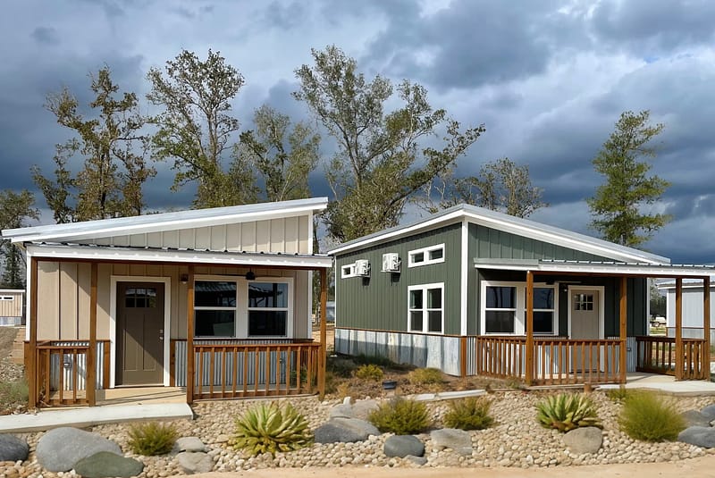 Two modern tiny houses with front porches sit side by side on a landscaped lot with rocks and small plants, under a cloudy sky and surrounded by trees.