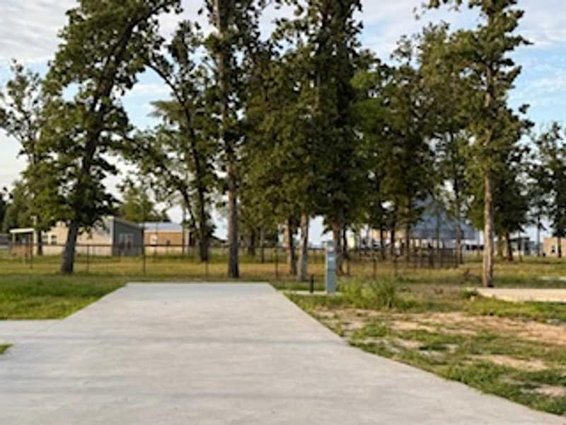 A concrete driveway leads to a fenced yard with several buildings and tall trees under a partly cloudy sky.