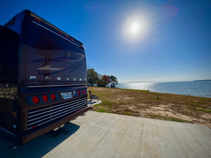A large black RV is parked on a concrete pad near a lake or bay under a bright, sunny sky with a few trees in the background.