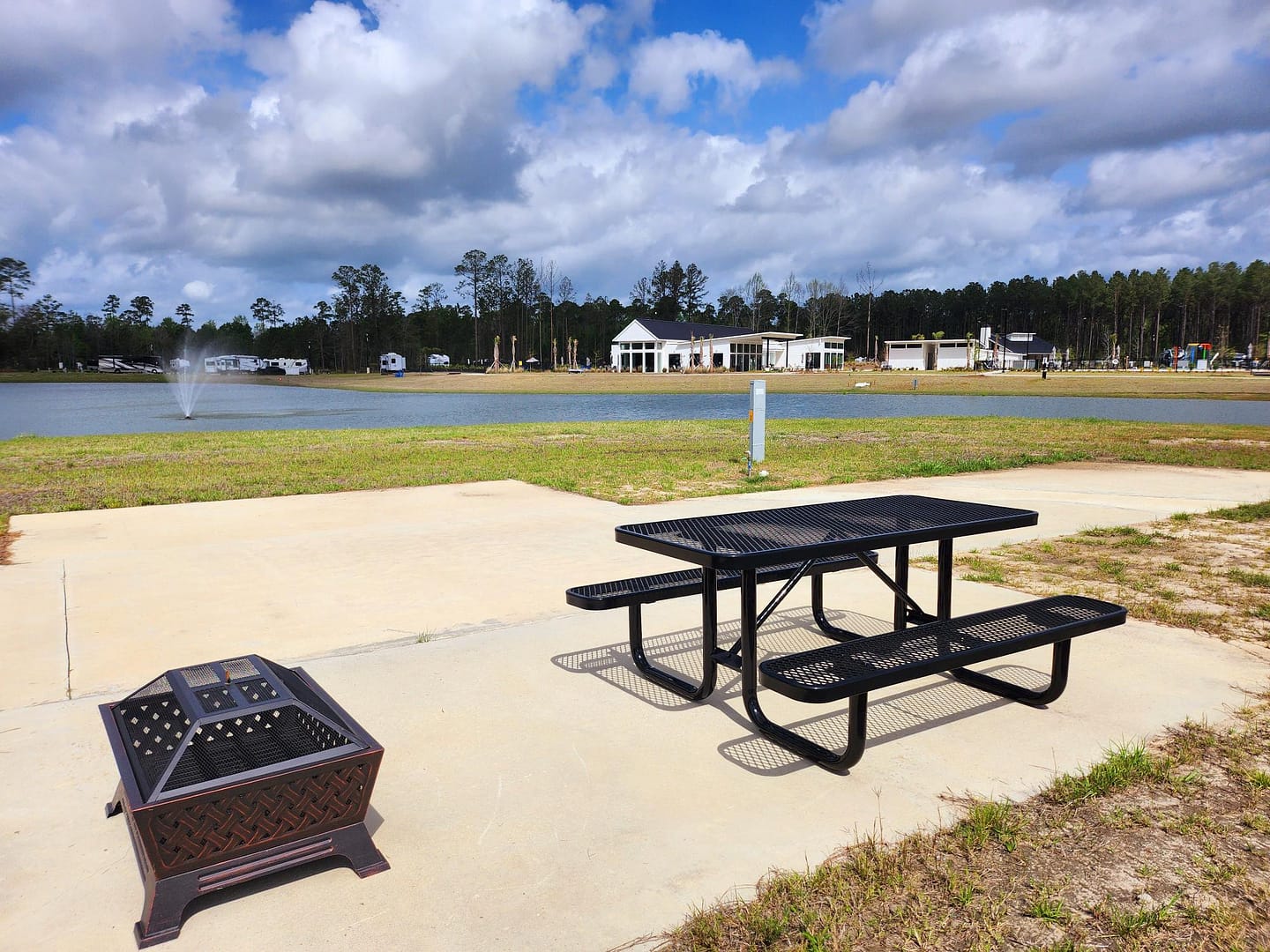 A black metal picnic table and fire pit sit on a concrete pad near a pond with a fountain, with buildings, trees, and RVs visible in the background under a partly cloudy sky.