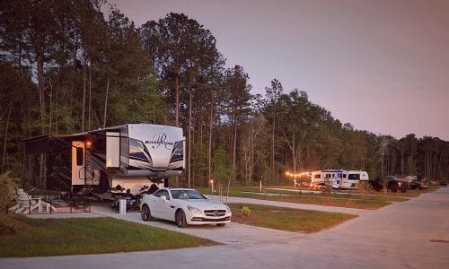 A white car is parked in front of a large RV at a campsite surrounded by trees, with other RVs and vehicles in the background.