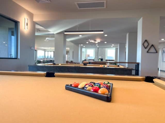 A close-up of a racked billiard set on a yellow pool table in a modern, well-lit game room with large windows and minimal decor.