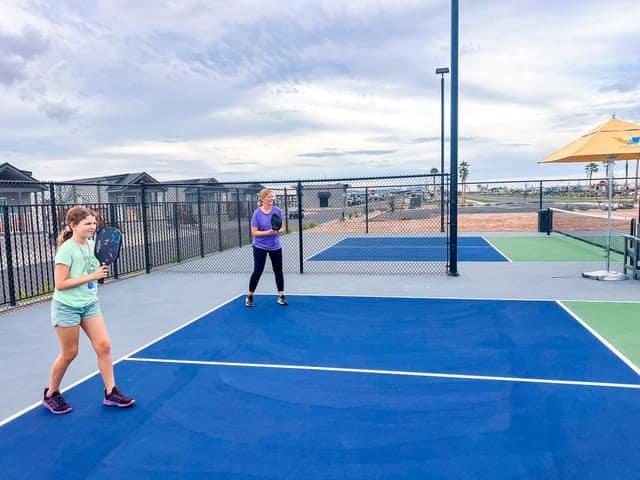 Two people are playing pickleball on an outdoor court with a chain-link fence and buildings in the background under a cloudy sky.