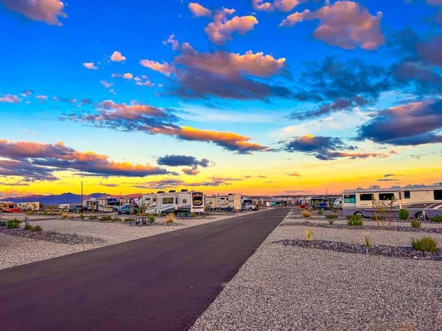 A paved road runs through an RV park at sunset, with parked RVs on both sides and a colorful sky with clouds overhead.