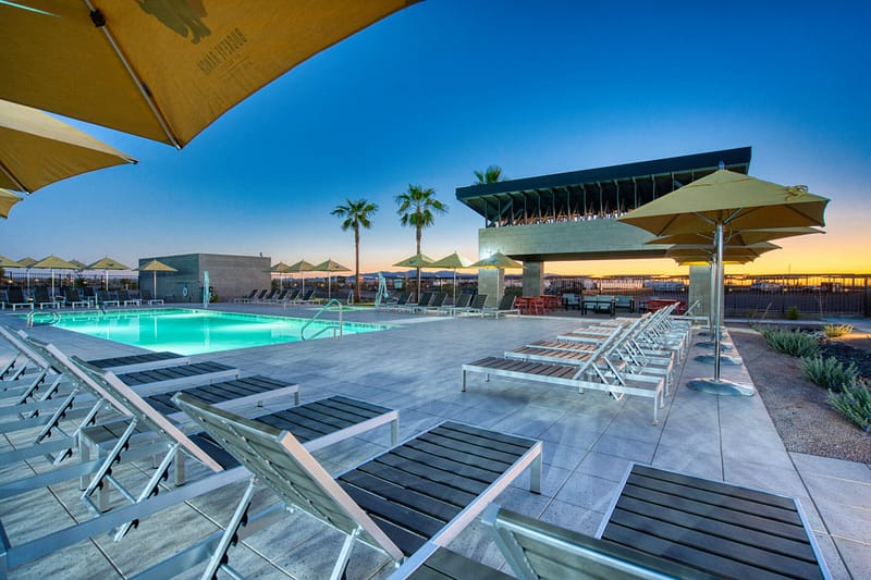 Outdoor pool area at sunset with lounge chairs, umbrellas, palm trees, and a modern building in the background.