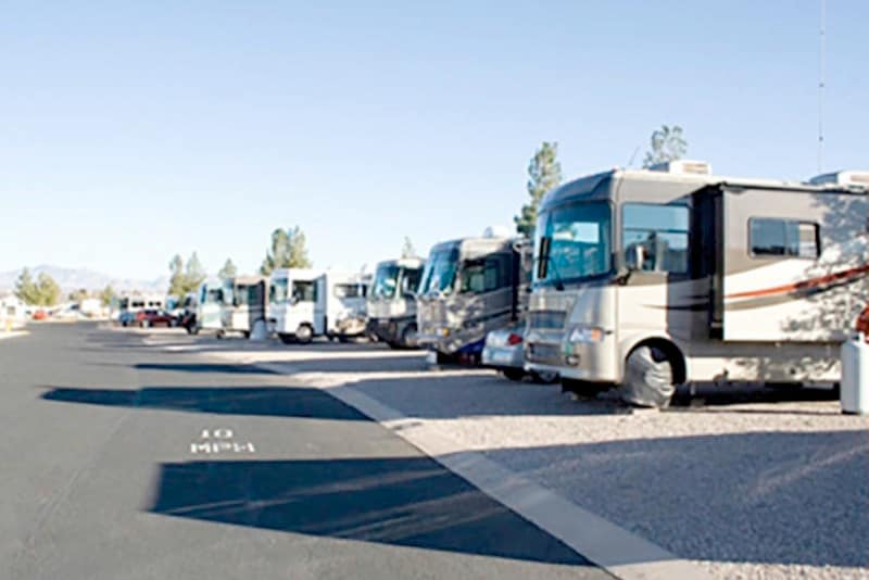 Several RVs are parked in designated spots along a paved road in an RV park, with clear skies and some trees in the background.