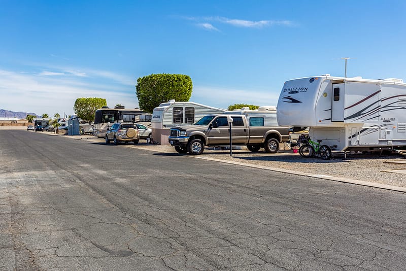 Several RVs and trucks are parked in designated spaces along a paved road in an RV park on a clear, sunny day.