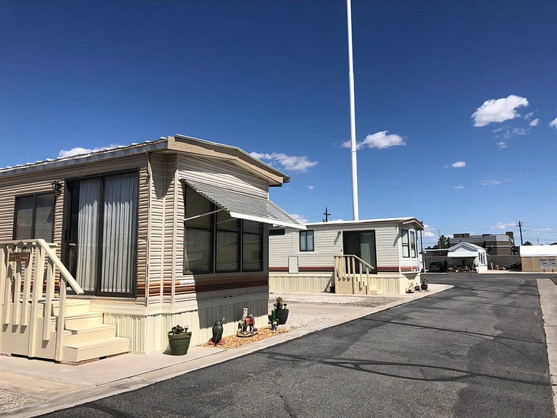 Two beige mobile homes with awnings are positioned along an asphalt road under a clear blue sky, with small plants near the entrances.