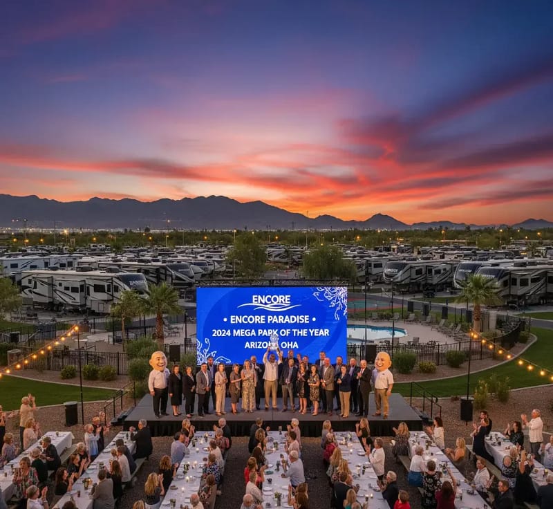 A group of people stands on an outdoor stage in front of a large screen reading "Encore Paradise 2024 Mega Park of the Year Arizona OHA," with RVs and mountains in the background at sunset.
